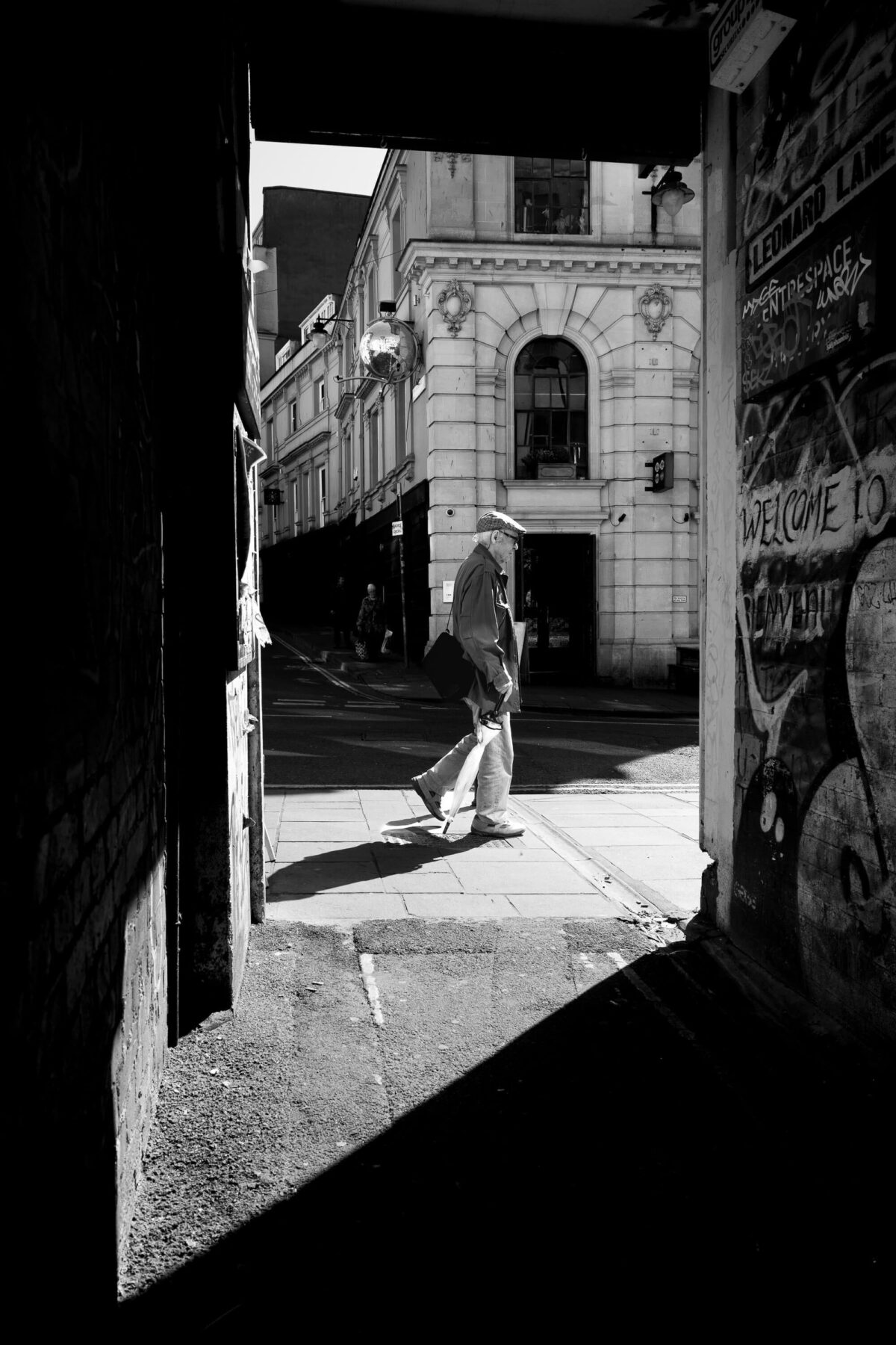 Elderly man in a flat cap walking through a sunlit Bristol passageway, candid street photography