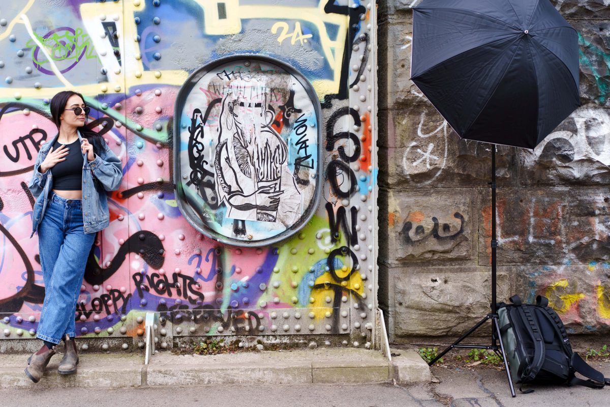 Tash leaning against a graffiti wall in a denim jacket, studio umbrella visible in the background