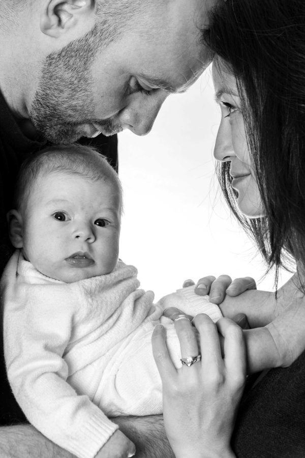 Mother, father, and baby look at each other. Baby pulling funny expression. White background.