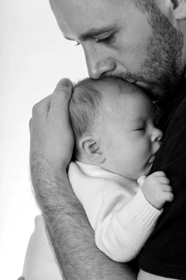 Ben gently pressing his lips to the top of baby Elidy's head in a tender kiss, white background