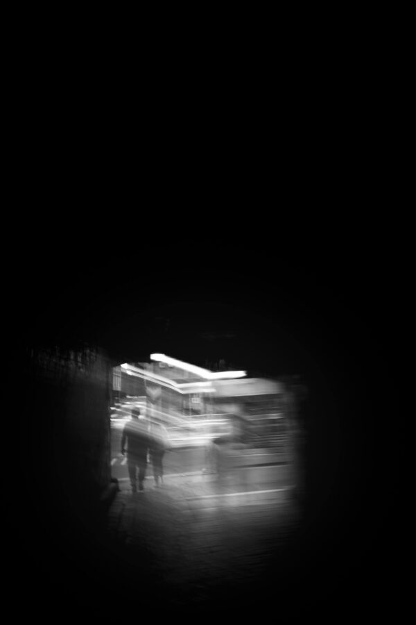 Long-exposure night street scene with bus light trails and a silhouetted pedestrian figure